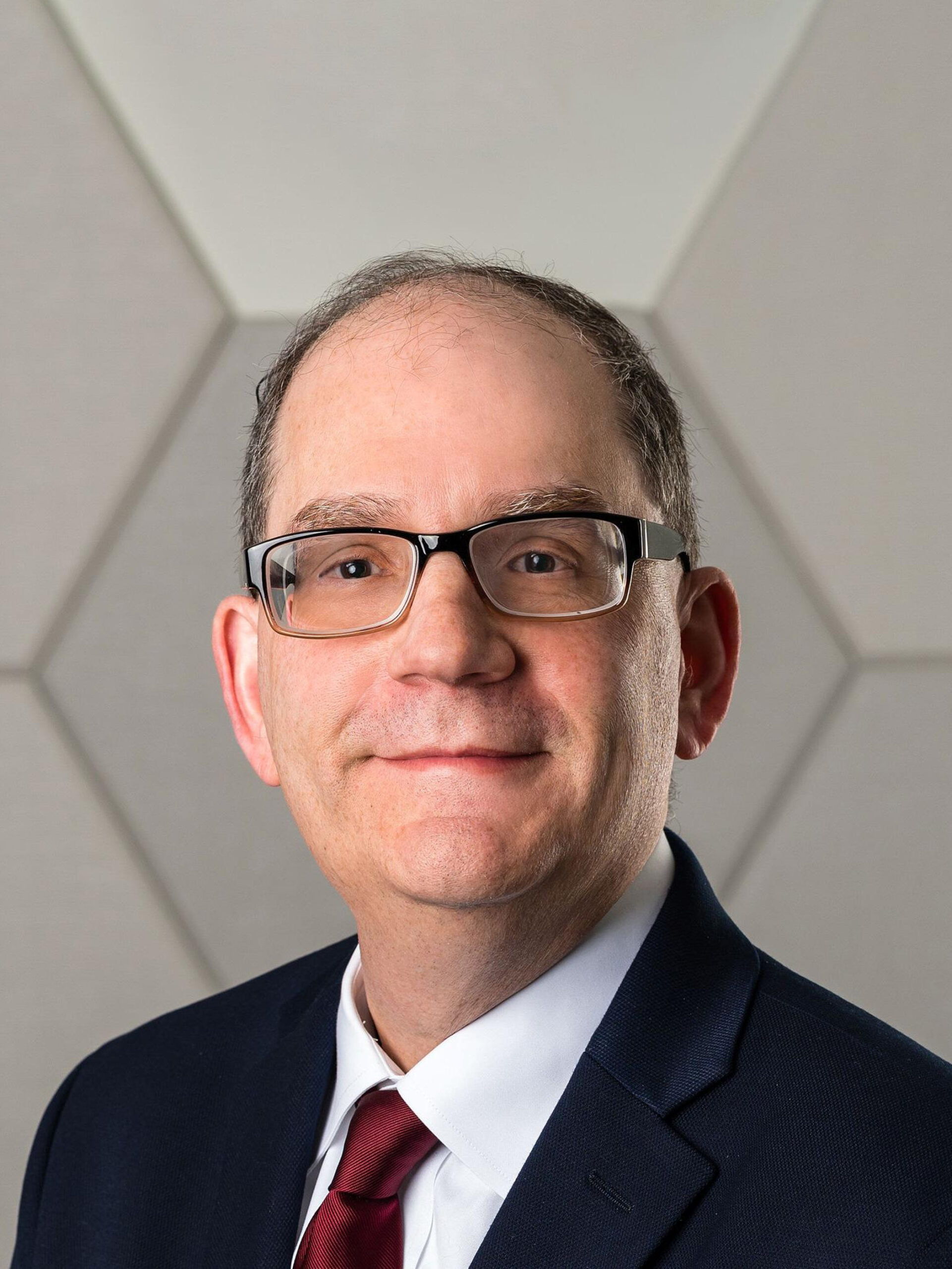 Man with glasses looks at the camera and smiles, wearing suit and red tie - Headshot of Dr. William Adelman