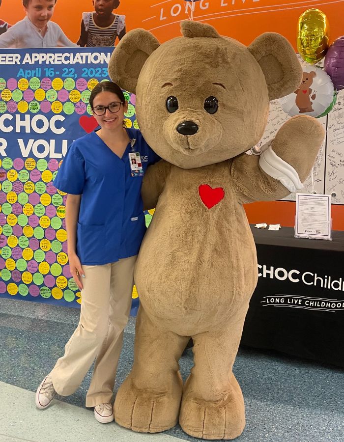 Smiling woman with dark hair and wearing a blue smock poses with a large plush teddy bear