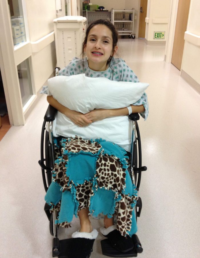 Girl with dark hair wearing a hospital gown and holding a pillow sits in wheelchair inside hospital 