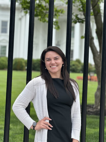Woman with brown hair wearing black dress and white sweater poses in front of the white house in washington dc