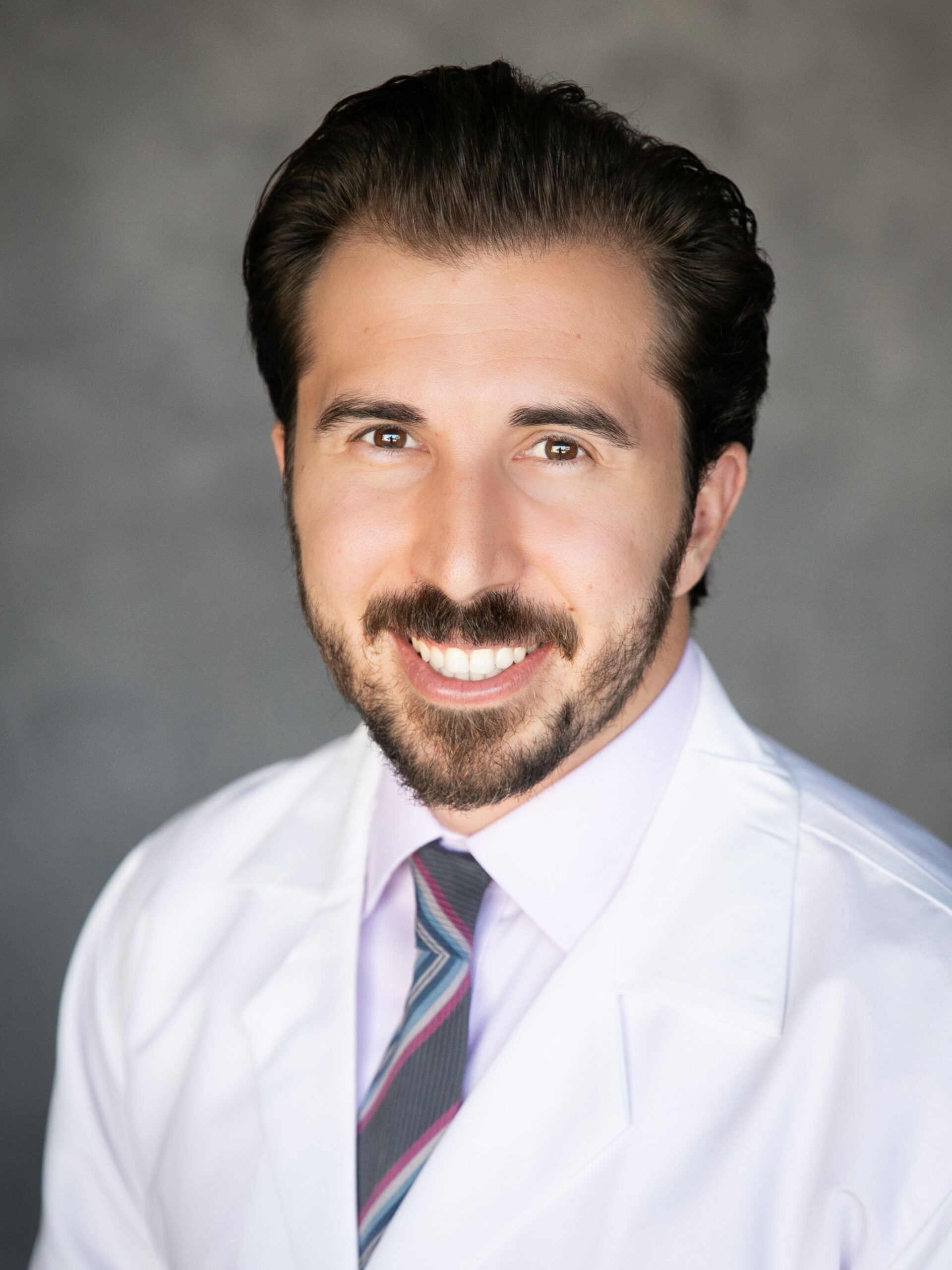 Man with dark hair and beard wearing a white doctors coat and tie