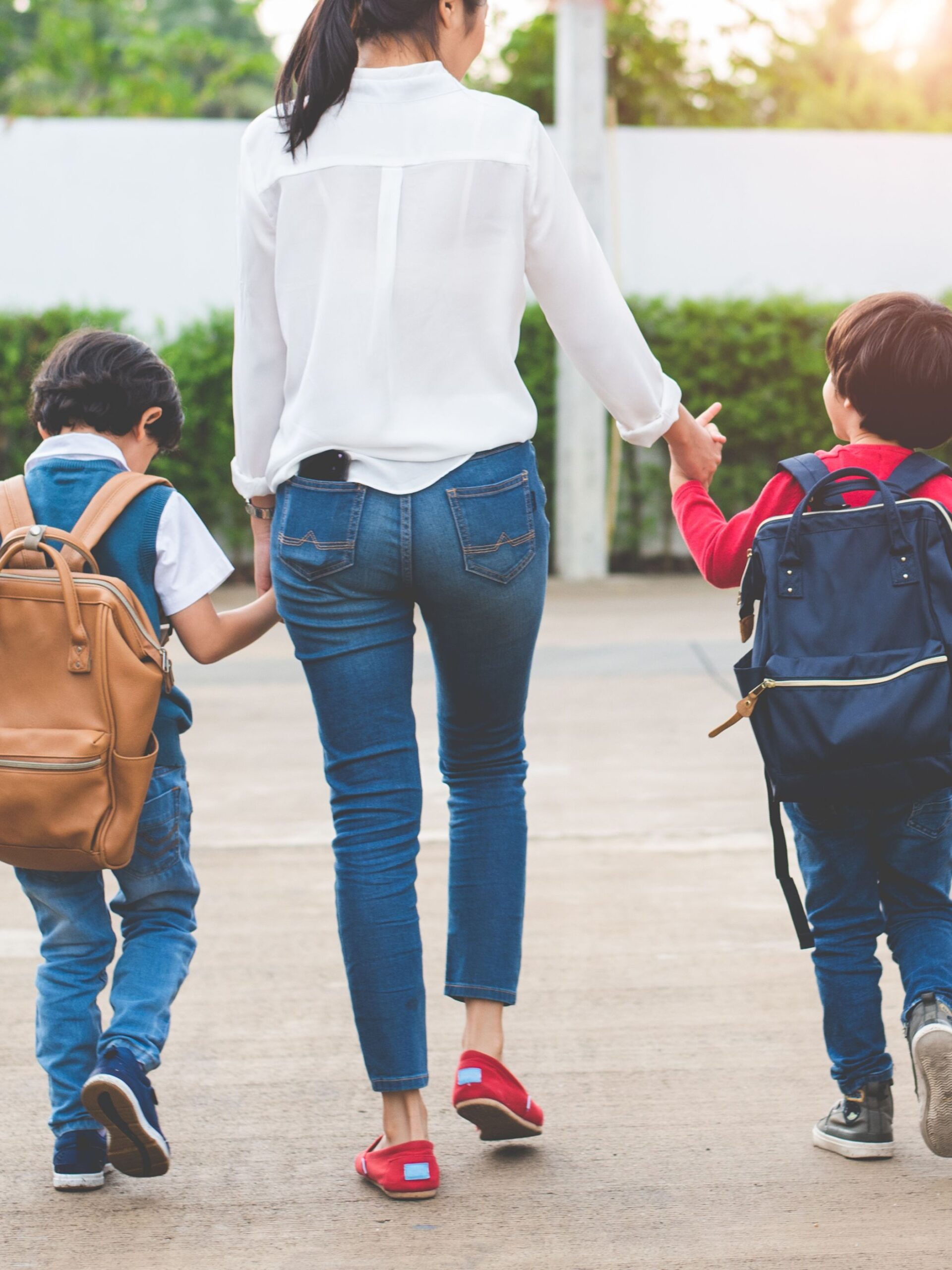 The back of a woman wearing a white blouse and blue jeans holding hands with two small kids wearing backpacks.