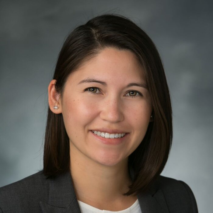 Professional headshot of a woman with shoulder-length dark hair wearing a dark blazer and white top.