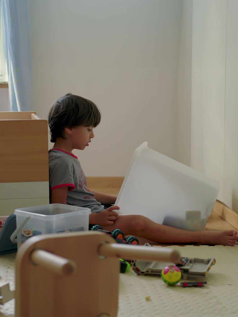 Young boy taking out toys from a plastic container, visibly upset and bored, sitting on the floor in his room with scattered toys and a bed nearby